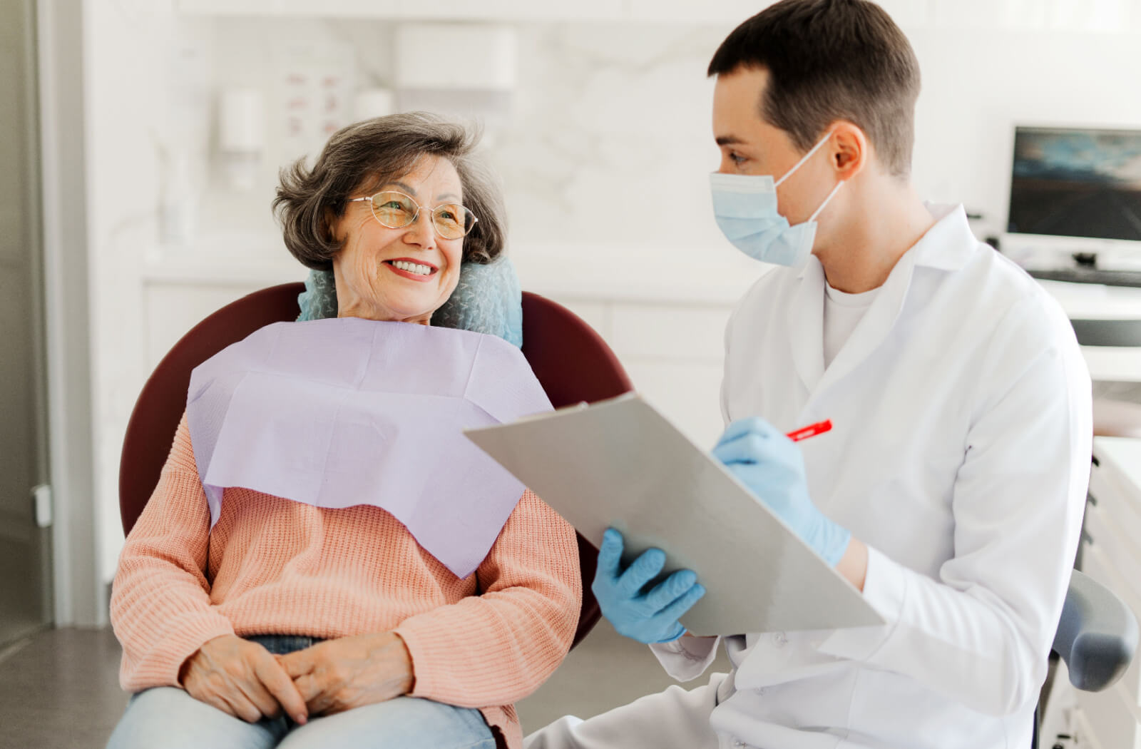 A dentist holds a clipboard and explains to a smiling older patient what's covered during their dental visit
