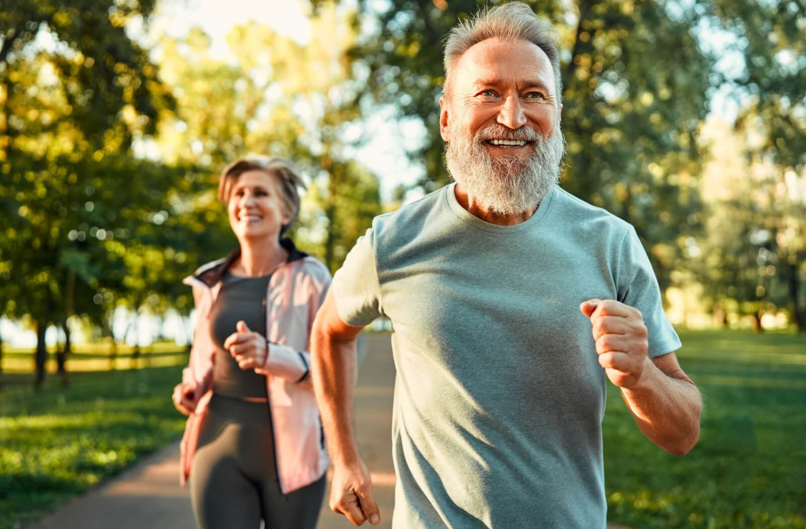 An older adult couple smiles while they jog through a local park, staying physically active to support their long-term memory