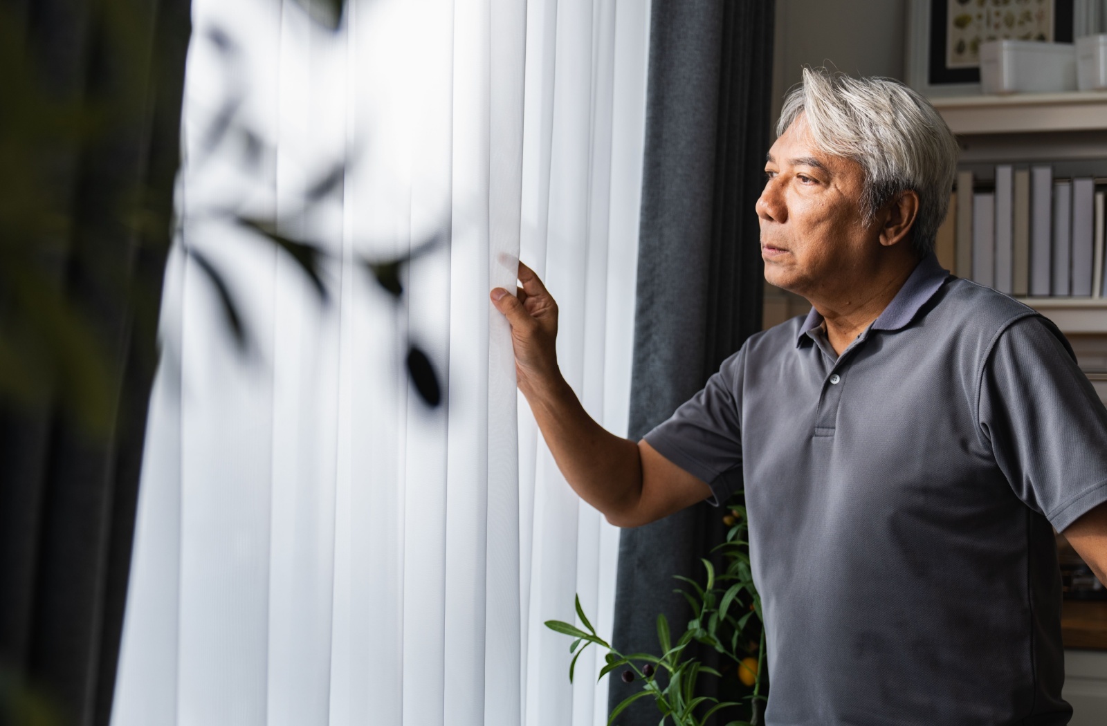 Older adult standing by a window pulling back the curtain with a thoughtful look