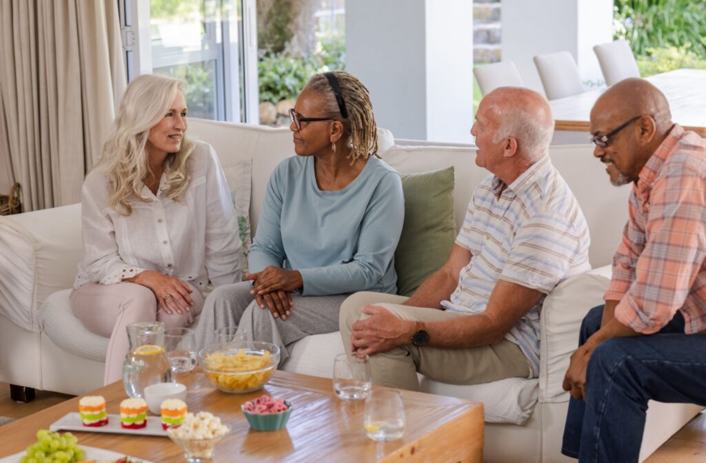 Group of older adults sitting together on a couch talking and sharing snacks in a bright living room